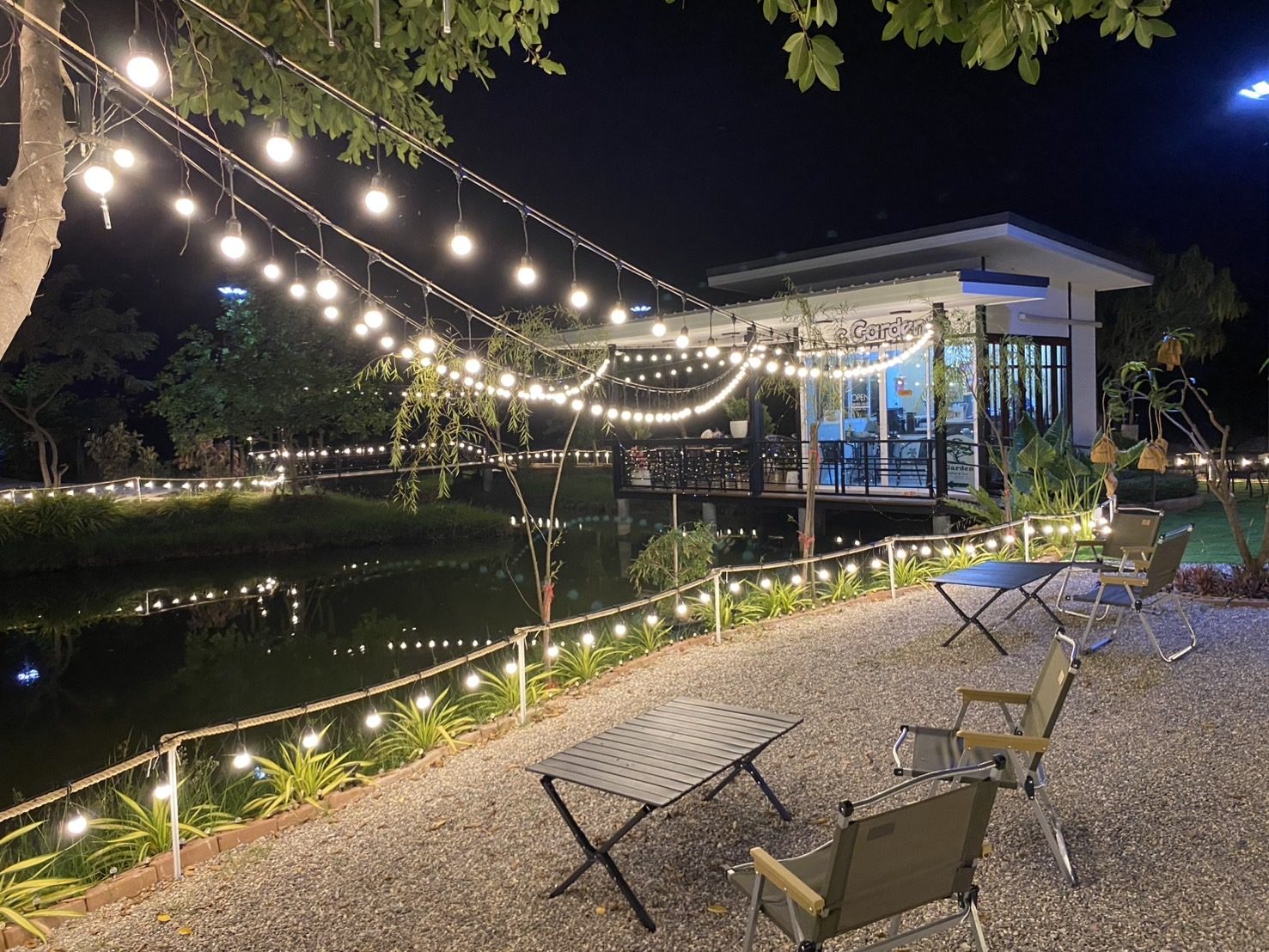 สวนริมสระของร้านยามค่ำ ไฟสายพาดทั่วสวน อาคารกระจกสว่างและเงาสะท้อนบนผิวน้ำ