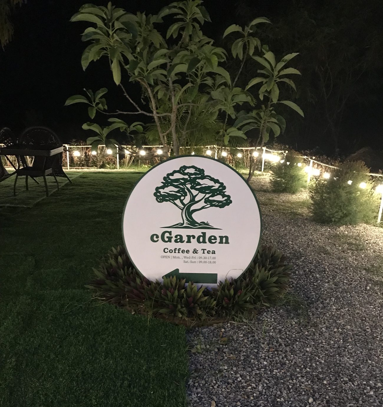 ป้ายกลมโลโก้ต้นไม้ cGarden Coffee & Tea หน้าร้านยามค่ำ มีไฟสายและสนามหญ้าโดยรอบ
